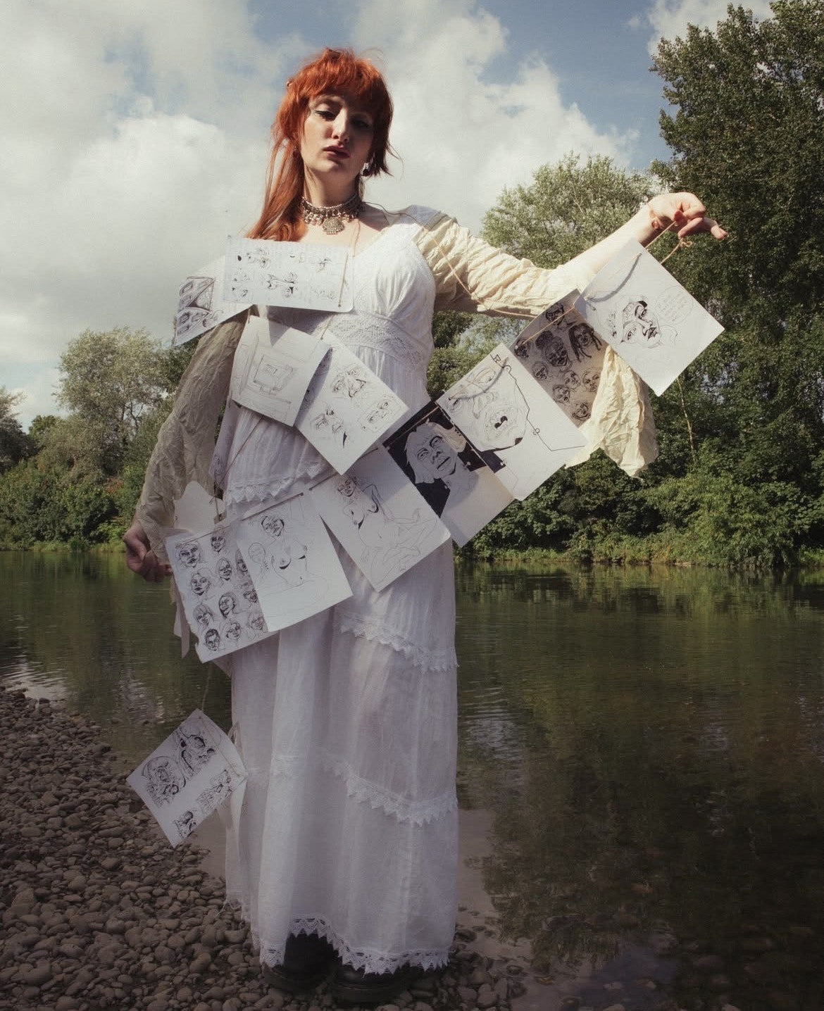 Person in a white dress holding sheets of paper with illustrations outdoors by a body of water.